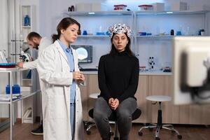 Stressed patient sitting on neurological chair with eeg headset, researcher examining health status writing on tablet. Medical scientist looking through microscope in background developing treatment. photo