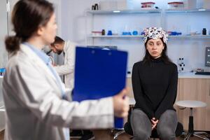 Medic researcher pointing at clipboard explaning treatment against brain disease to patient with eeg headset. Woman sitting in neurological scientific laboratory treating nervous system dysfunctions. photo
