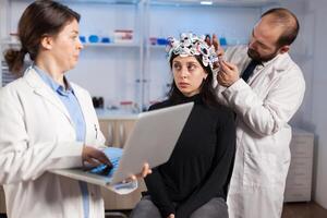 Expert medic in neuroscience developing brain experiment holding laptop explaning to woman side effects of nervous system treamtment. Team of scientists working in neurological research laboratory. photo