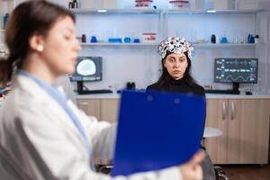 Neuroscience doctor holding clipboard showing treatment against brain disease to patient with eeg headset. Woman sitting in neurological scientific laboratory treating nervous system dysfunctions. photo