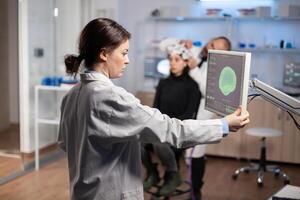Neurologist doctor analysing brain scan on monitor display in modern research laboratory, developing a cure for nervous system diagnosis. Woman patient wearing headset with sensors photo
