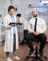 Specialist doctor in neuroscience taking notes on clipboard while testing brain functions of man using eeg headset, treating nervous system dysfunctions in moden laboratory. photo