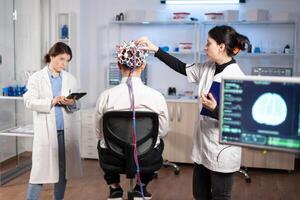 Back view of man patient wearing performant brainwave scanning headset sitting in neurological research laboratory while medical researcher adjusting it, examining nervous system typing on tablet. photo