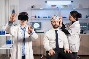 Neuroscientist developing diagnosis wearing virtual reality headset in medicine laboratory, assistant adjusting examining sensors on patient sitting on chair. Medicine lab with modern technology. photo