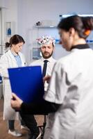 Researcher neurologist doctor asking man's illness symptoms looking at clipboard before brain scan with brainwave scanning headset. Scientist analyzing health status, nervous system, tomography scan. photo