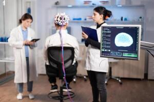 Man patient wearing performant eeg headset scanning brain electrical activity in neurological research laboratory while medical researcher controlling it, examining nervous system, nurse taking notes photo
