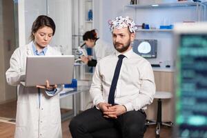 Medic in neuroscience working in neurological research laboratory developing brain experiment holding laptop explaning to man brainwave scanning headset side effects of nervous system treamtment. photo