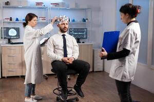 Ill man listening neurological doctor looking at clipboard wearing high tech headset scanning brain electrical activity. Patient sitting in scientific laboratory treating nervous system dysfunctions photo
