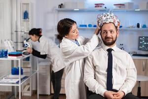 Woman researcher putting brainwave scanning headset to patient developing experiments using brain study lab monitors. Man siting on chair while scientist adjusting device examining nervous system photo