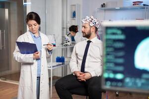 Medical doctor talking about symptoms of disease during investigations using high tech brainwave scanning headset. Researcher analysing health status of man patient, brain functions, nervous system. photo