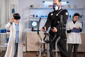 Scientist doctor in sport science laboratory wearing virtual reality goggles while athlete running, with electrodes attached on body monitoring physical endurancewhile ekg scan runs on computer screen. photo