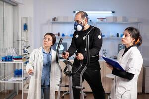 Team of scientist monitoring vo2, cardio ekg of performance athlete, running during cross training with electrodes for measuring attached to body in laboratory. photo