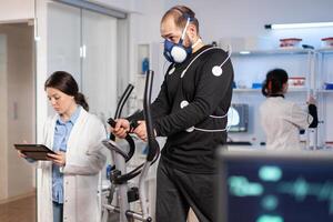 Performance athlete running on cross trainer with electrodes attached on body and mask in testing lab, scientist doctor monitoring heart rate looking at tablet pc. photo