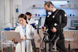 Man athlete running on cross trainer with electrodes attached to his body and mask. Physician usinglaptop and controls EKG data showing on laboratory monitors, discussing with patient. photo