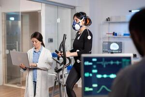 Team of medial researchers monitoring vo2 of woman performance sports wearing mask running. Lab science doctor measuring endurance using tablet while ekg scan runs on computer screen in laboratory. photo