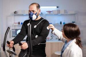 Man athlete with mask running on cross trainer after medical researcher attached electrodes to her body. Physician using computer to controls EKG data showing on laboratory monitors, VO2 max test. photo