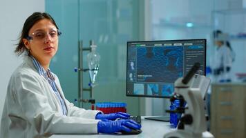 Portrait of tired scientist trying to smile at camera sitting in modern equipped laboratory typing on computer. Chemist examining virus evolution using high tech for scientific research, vaccine photo