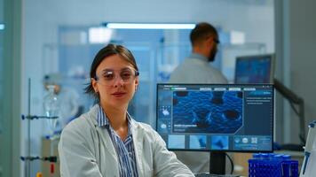 Chemist with lab coat sitting in laboratory looking at camera smiling, while colleague working on computer in background. Scientist examining virus evolution using high tech for scientific research photo