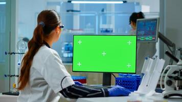 Woman researcher typing at computer with green screen mockup display, template in applied science laboratory. Engineer conducting experiments in background, examining vaccine evolution using high tech photo