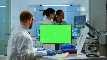 Man researcher looking at chroma key display in modern equipped lab typing on computer. Team of microbiologists doing vaccine research writing on device with green screen, isolated, mockup display. photo