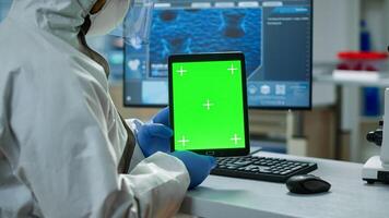 Chemist man wearing protection suit holding tablet with green mockup in modern equipped lab. Team of microbiologists doing vaccine research writing on device with chroma key, isolated display. photo