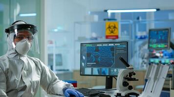 Tired laboratory doctor with overall suit working at computer and looking at camera. Scientist examining virus evolution using high tech, chemistry tools for scientific research, vaccine development photo