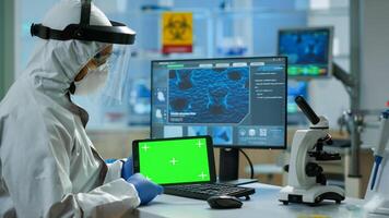 Doctor with coverall working at tablet with green screen in modern equipped lab. Team of microbiologists doing vaccine research writing on device with chroma key, isolated, mockup display. photo