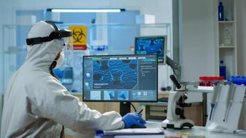 Scientist in protection suit typing on pc working in medical laboratory, writing on clipboard, developing inovative drugs against new virus. Doctor using high tech, chemistry tools for researching photo