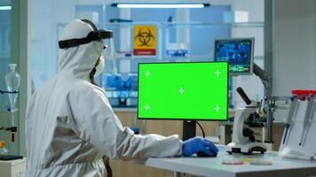 Scientist man wearing protection suit typing on computer with green mockup in modern equipped lab. Team of microbiologists doing vaccine research writing on device with chroma key, isolated display. photo