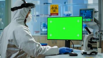 Man researcher in coverall looking at chroma key computer in modern equipped lab. Team of microbiologists doing vaccine research writing on device with green screen, isolated, mockup display. photo