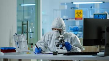 Biologist in ppe suit working on microscope and writing information on clipboard in equipped laboratory. Chemist examining vaccine evolution using high tech researching diagnosis against covid19 virus photo