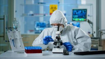 Scientist in ppe suit making adjustments and looking through laboratory microscope, typing on pc. Chemist in coverall working with various bacteria, tissue blood samples for antibiotics research photo