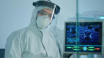Microbiologist in ppe suit standing in laboratory looking at camera behind the glass wall in equipped lab. Doctor examining virus evolution using high tech and chemistry tools for vaccine development photo