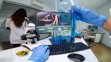 FPV of doctor chemist holding a flask of blue solution looking at it and typing on computer medical results. Scientists examining vaccine evolution in medical lab using high tech chemistry tools photo
