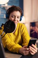 Happy blogger reading messages of fans using smartphone sitting in home podcast studio during livestreaming, recording. Online show On-air production internet broadcast host streaming live content. photo