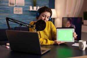 Influencer holding tablet with green screen and speaking into microphone during podcast in home studio. On-air production internet broadcast host streaming live content using chroma key mockup display. photo