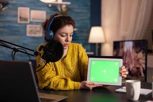 Vlogger looking at laptop and talking about tablet with chroma key desktop. On-air production internet broadcast host streaming live content using mochup, green screen, izolated desktop. photo