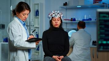 Stressed patient sitting on neurological chair with eeg headset, researcher examining health status writing on tablet. Medical scientist looking through microscope in background developing treatment photo