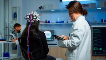 Back view of woman patient wearing performant eeg headset sitting on chair in neurological research laboratory while medical researcher adjusting it, examining nervous system typing on tablet. photo