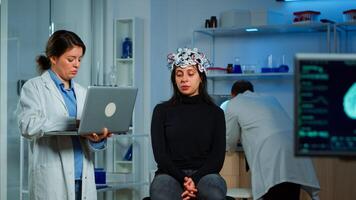 Specialist neurologist doctor taking notes on laptop asking patient's symptoms adjusting high tech eeg headset. Doctor researcher controlling EEG headset analyzing brain functions and health status photo