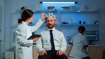 Young man wearing brainwave scanning headset discussing with neurological researcher about symptoms of disease. Medical scientist writing informations of brain scan on tablet working in laboratory photo