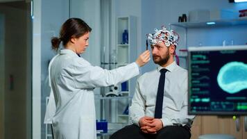 Man sitting on neurological chair with brainwave scanning headset while researcher examining health status writing on clipboard. Doctors working in brain study lab with monitors showing EEG reading. photo
