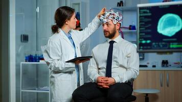 Man sitting on neurological chair with brainwave scanning headset, researcher doctor tracking evolution of patient's brain using tablet, adjusting eeg device. Scientist examining nervous system. photo