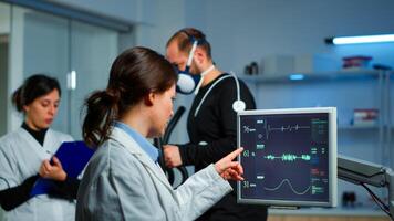 Muscular athlete with mask running on cross trainer in medical laboratory for monitoring his performance while ekg scan runs on computer screen and team of doctor researcher examining results photo