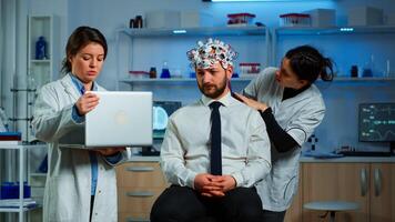 Medic in neuroscience working in neurological research laboratory developing brain experiment holding laptop explaning to man brainwave scanning headset side effects of nervous system treamtment. photo