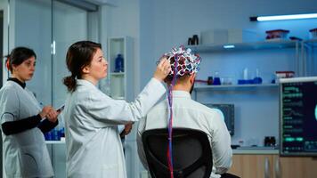 Back view of man patient wearing performant brainwave scanning headset sitting in neurological research laboratory while medical researcher adjusting it, examining nervous system typing on tablet. photo