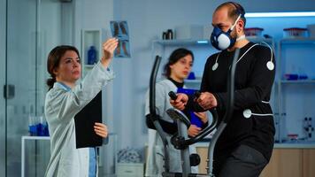 Team of doctor researchers monitoring endurance of man performance sports wearing mask running cross trainer. Lab science doctor measuring vo2 of sportsman, looking at x-ray explaning health status photo