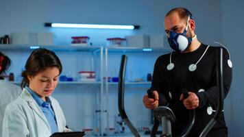 Doctor science typing on tablet while measuring sportsman endurance using body sensors and mask monitoring cardiac rhythm. Man running on cross trainer in sports lab, specialists supervise exercise photo