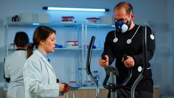 Athlete with mask and electrodes attached on body running on cross trainer in science sport lab monitoring health status. Medical researcher measuring his performance cardio respiratory using laptop photo