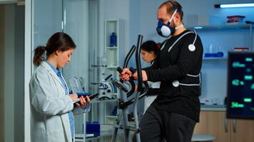 Science sport doctor asking patient about his health while sportsman running on cross trainer with mask and electrodes attached on body monitoring vo2. Woman researcher measuring endurance of patient. photo